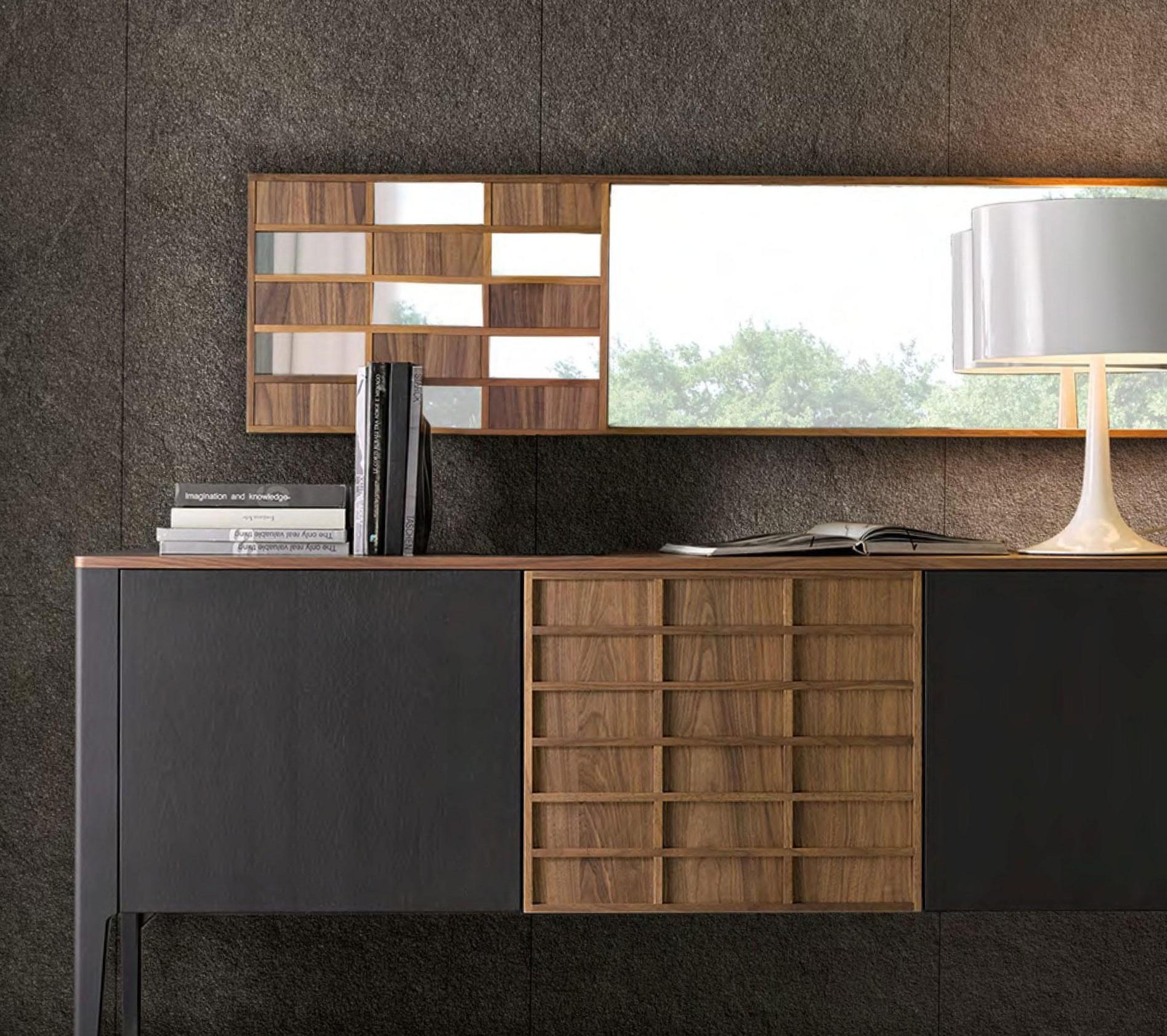 A modern Italian mirror with a geometric walnut wood frame featuring a grid pattern of alternating reflective panels, mounted above a matching black and walnut sideboard styled with stacked books and a white table lamp.